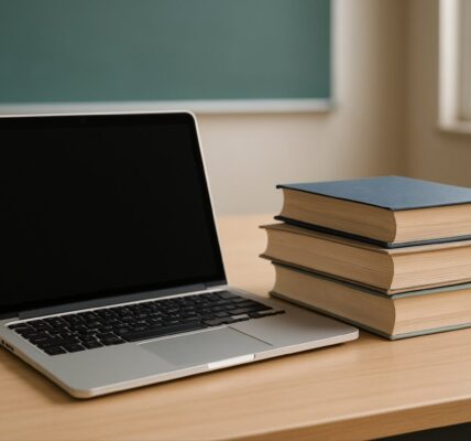 A laptop next to stacked books on a wooden desk against a classroom backdrop